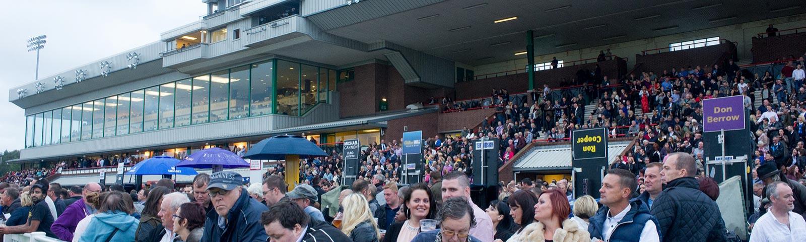 Crowds watching racing from the grandstand, with more people gathered around bookmakers.